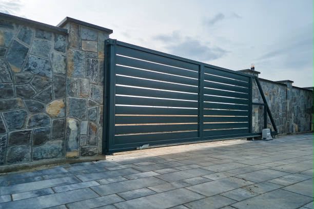 A dark grey horizontal-slat sliding gate, closed between stone-clad piers, across a paved driveway.
